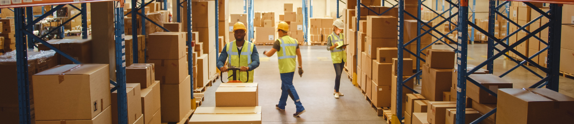 Interior of a spacious warehouse with high ceilings and bright lighting. Tall shelves stocked with neatly organized boxes line the aisle. Three workers wearing safety vests and helmets are handling and moving boxes, with one using a cart for efficient transport.