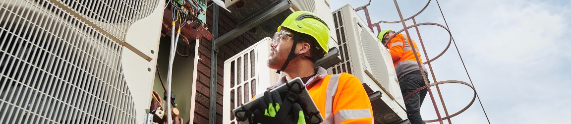 A HVAC technician in a high-visibility vest and safety helmet is working on large air conditioning units, using a digital tablet to inspect or perform maintenance. The industrial setting suggests a significant cooling requirement for a facility.