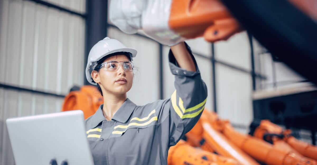 Young woman in safety goggles and hat inspects an orange robotic arm to determine the criticality of factory assets