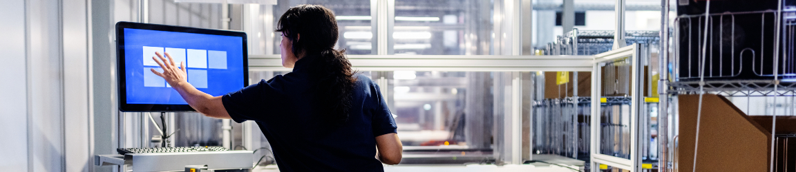 A young woman in a distribution warehouse is updating package details on a computer. She is focused on the screen, with shelves of packages and boxes in the background, indicating a busy and organized workspace. She is an example of how automation is changing warehouse work. 