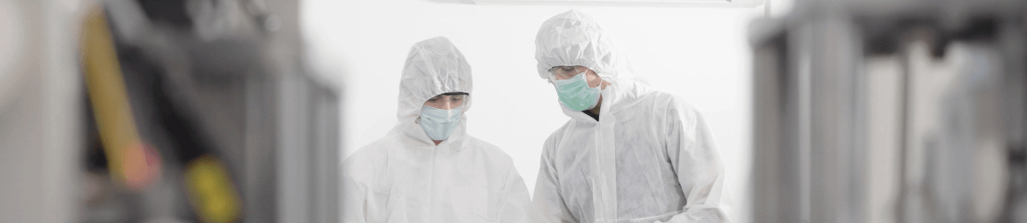 Two workers in a cleanroom wear white coveralls, gloves, facemasks and clear safety glasses as they review information on a tablet.