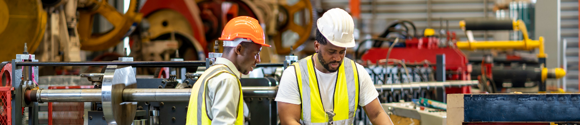 A team of industrial workers, wearing safety helmets and reflective vests, is checking the setup value of a metal sheet roll forming machine using an automated monitor inside a factory. The environment is well-lit, with various industrial equipment visible in the background, emphasizing a focus on safety and precision in their work.