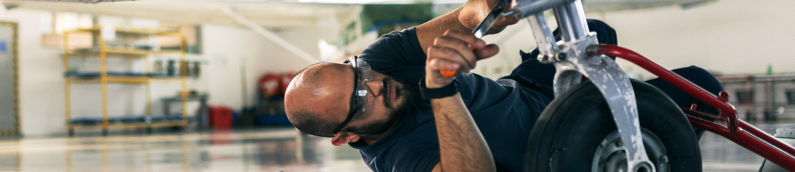 An aviation aircraft mechanic is servicing a plane in a well-lit workshop. The mechanic, wearing a blue uniform and safety glasses, is focused on inspecting the aircraft’s engine. Various tools and equipment are visible in the background, highlighting the technical environment of the maintenance facility.