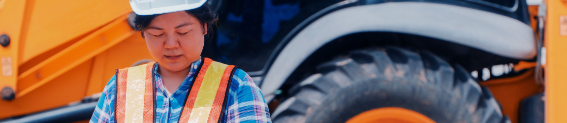 A heavy equipment operator wearing a white safety helmet stands confidently in front of a backhoe and construction site, holding a tablet, she is leveraging the latest technology in her work. The heavy equipment operator is dressed in a high-visibility vest and safety gear, symbolizing her role in overseeing the construction project.