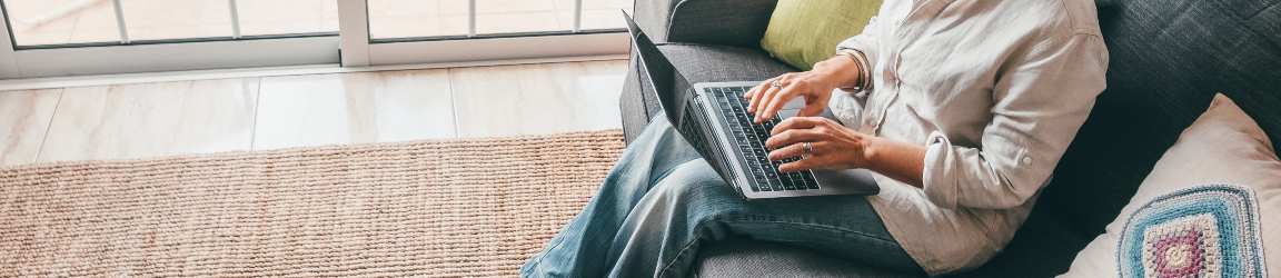 A smiling woman with curly hair sitting on a couch at home, using a laptop. She is searching for a job while wearing a grey shirt, blue pants and tan slippers. 