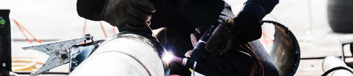 A pipefitter perches over a metal pipe while welding the end closest to them. The welder wears a long-sleeve dark blue shirt and dark grey gloves. They are also wearing a tan and grey welding hood. 